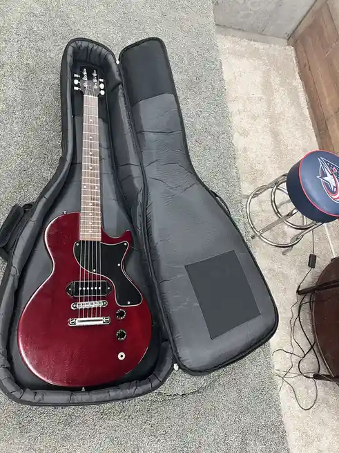 A red electric guitar rests inside an open black padded case on a gray carpet, with a blue and red bar stool and tangled…
