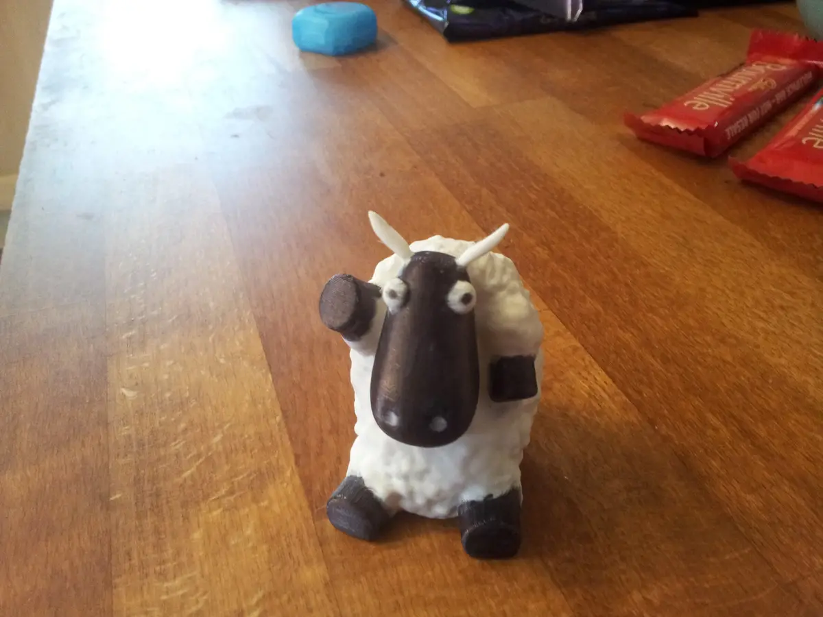A small, handmade figurine of a sheep with a dark brown face, horns, and hooves, sitting on a wooden table. The sheep is…