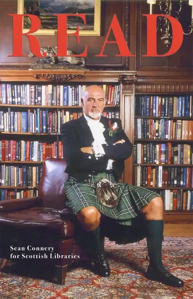 A man in a green kilt and formal jacket sits confidently in a leather chair in front of bookshelves filled with books. Large…