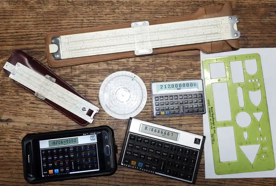A flat lay of vintage and modern calculators and slide rules on a wooden surface, including a slide rule in a leather case…