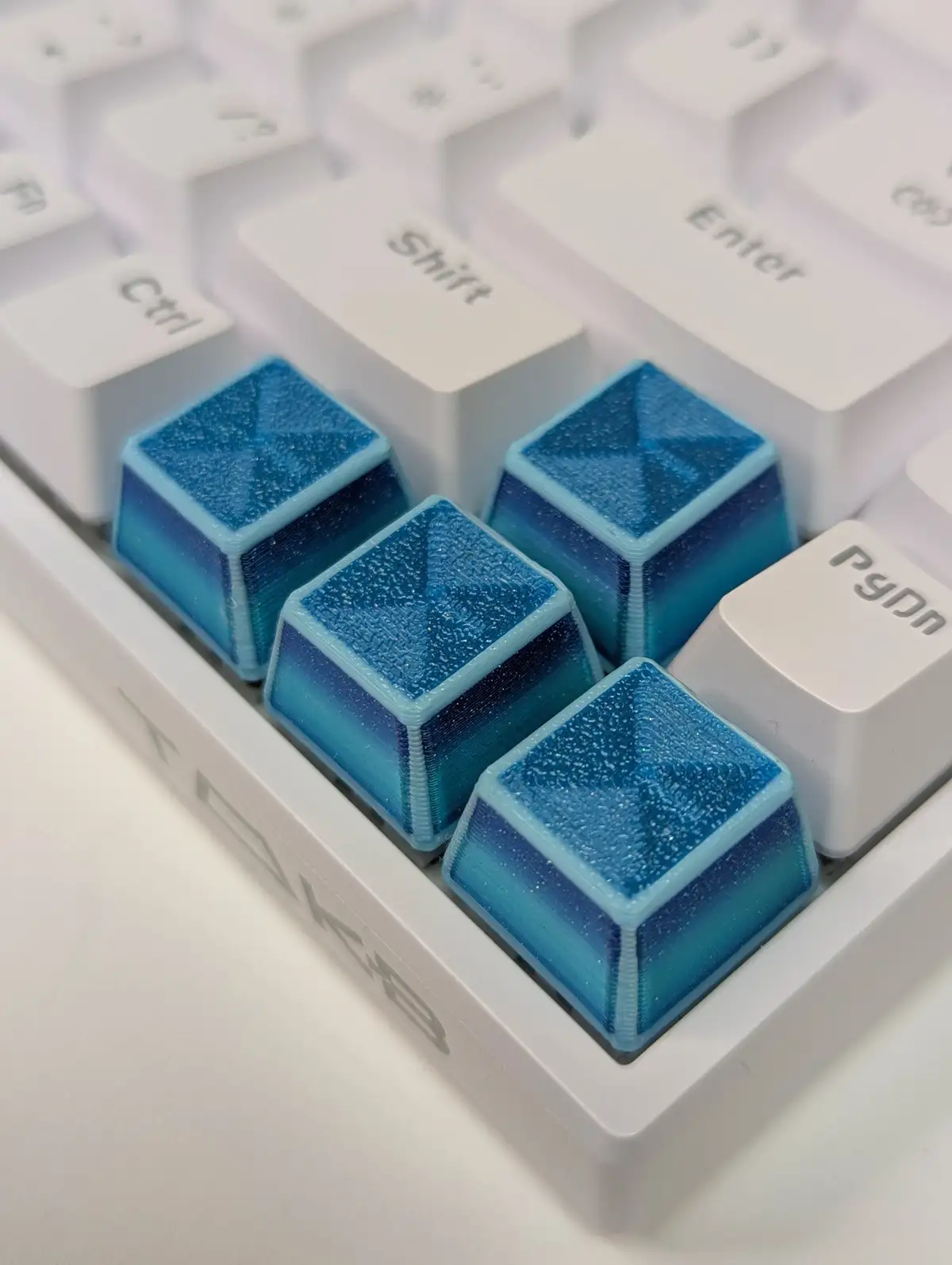 A close-up photo of a white mechanical keyboard with four custom blue gradient keycaps arranged in a cluster, next to a…