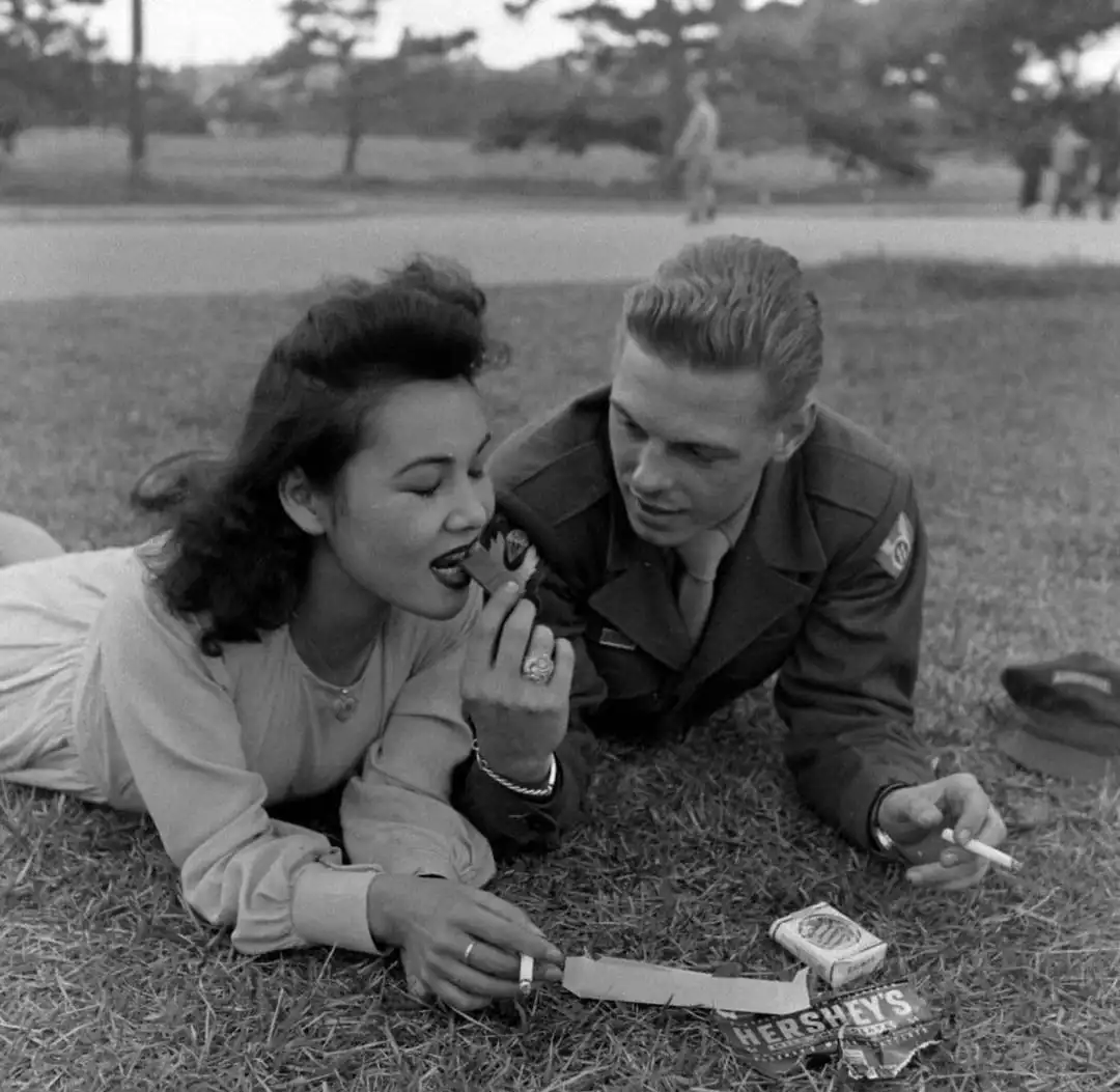 A black-and-white photo shows a woman and a man in a military uniform lying on grass, sharing a Hershey's chocolate bar. The…