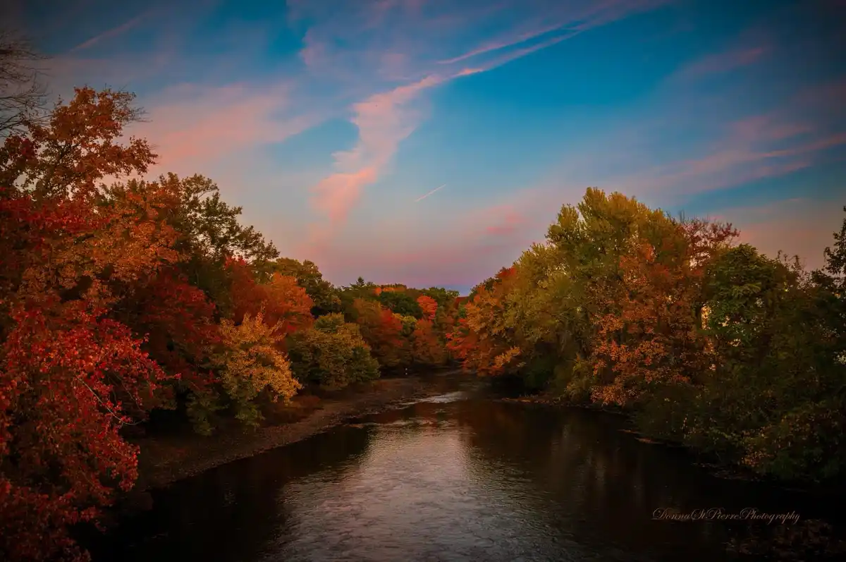 A serene autumn landscape featuring a river flanked by trees with vibrant red, orange, and yellow leaves under a twilight…