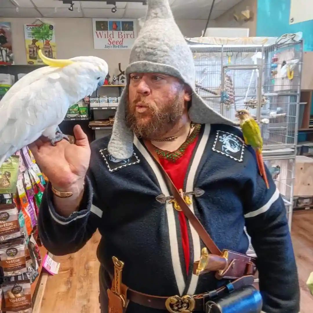 A man dressed in fantasy-themed clothing, including a tall grey hat and a dark blue tunic with white trim, stands inside a…