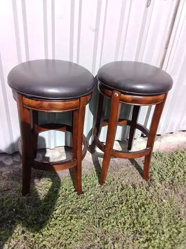 Two identical wooden bar stools with dark brown leather seats stand side by side on a patch of grass in front of a…