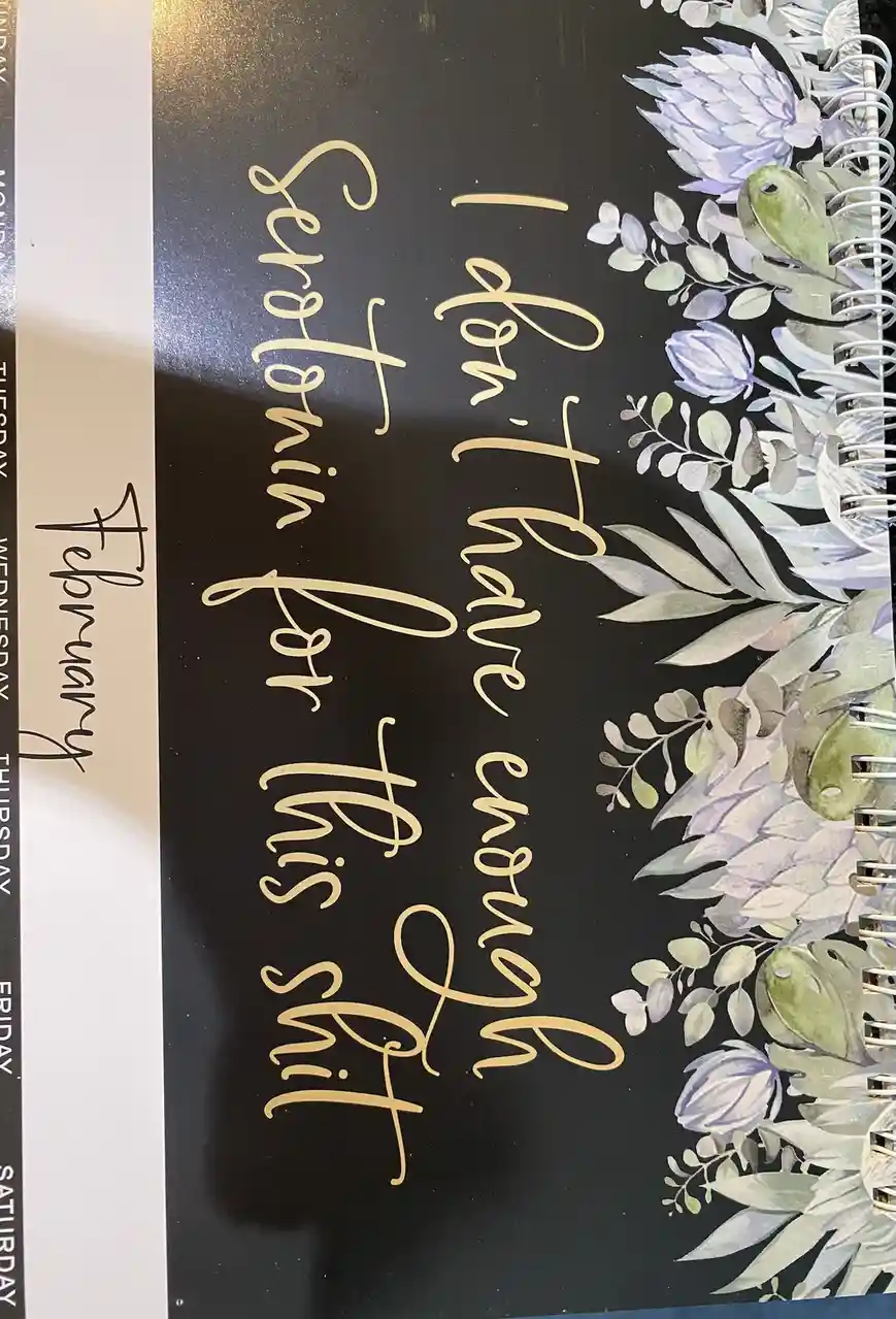 A close-up of a black spiral-bound planner with gold cursive text reading 'I don't have enough Serotonin for this shit' and…