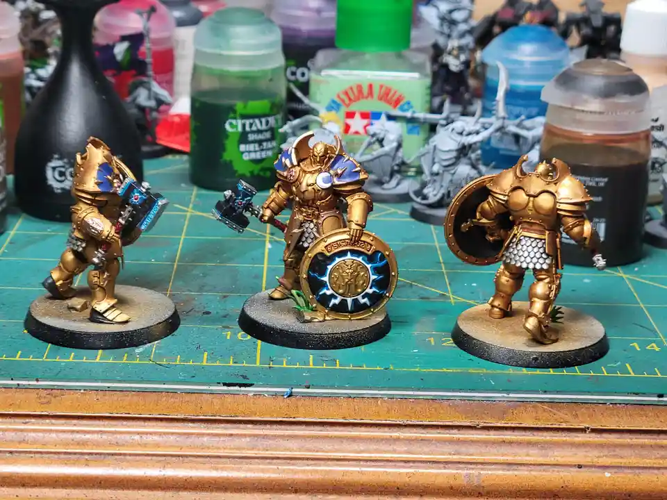 Three intricately painted fantasy miniatures, resembling heavily armored warriors with gold-colored armor, stand on circular…