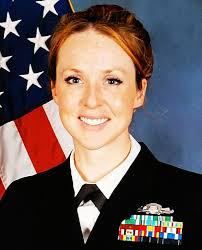A smiling woman in a dark military uniform with medals on her chest stands in front of an American flag. The overlaid text…