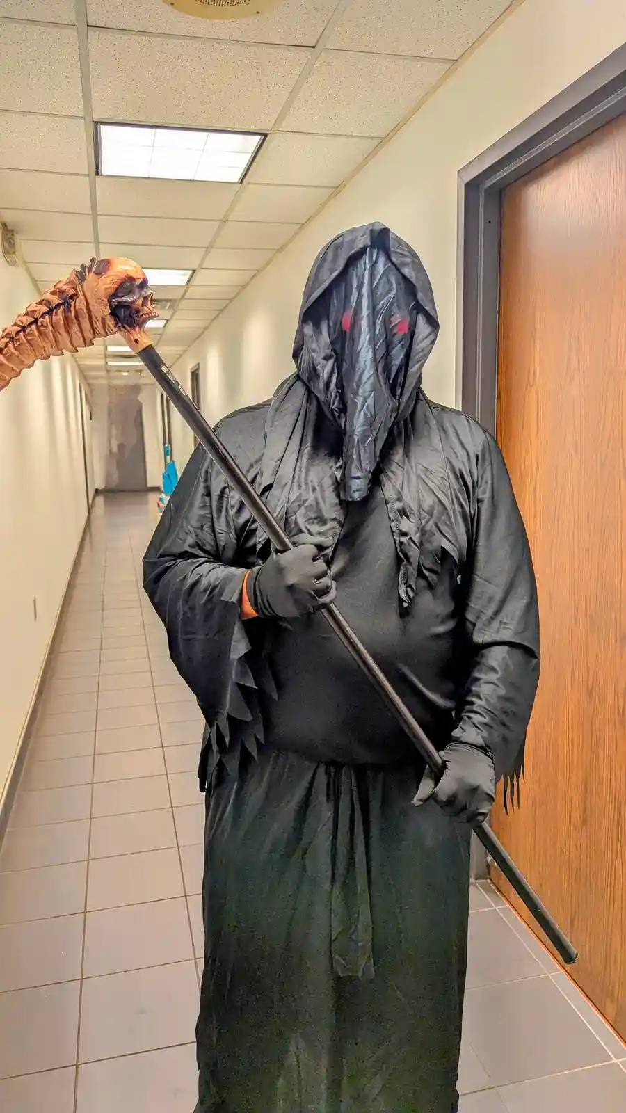 A person in a black hooded robe with a long, narrow face mask featuring red glowing eyes stands in an office hallway holding…