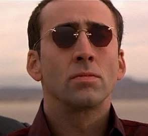 A close-up of actor Nicolas Cage wearing dark sunglasses and a maroon collared shirt, looking intensely forward with a…