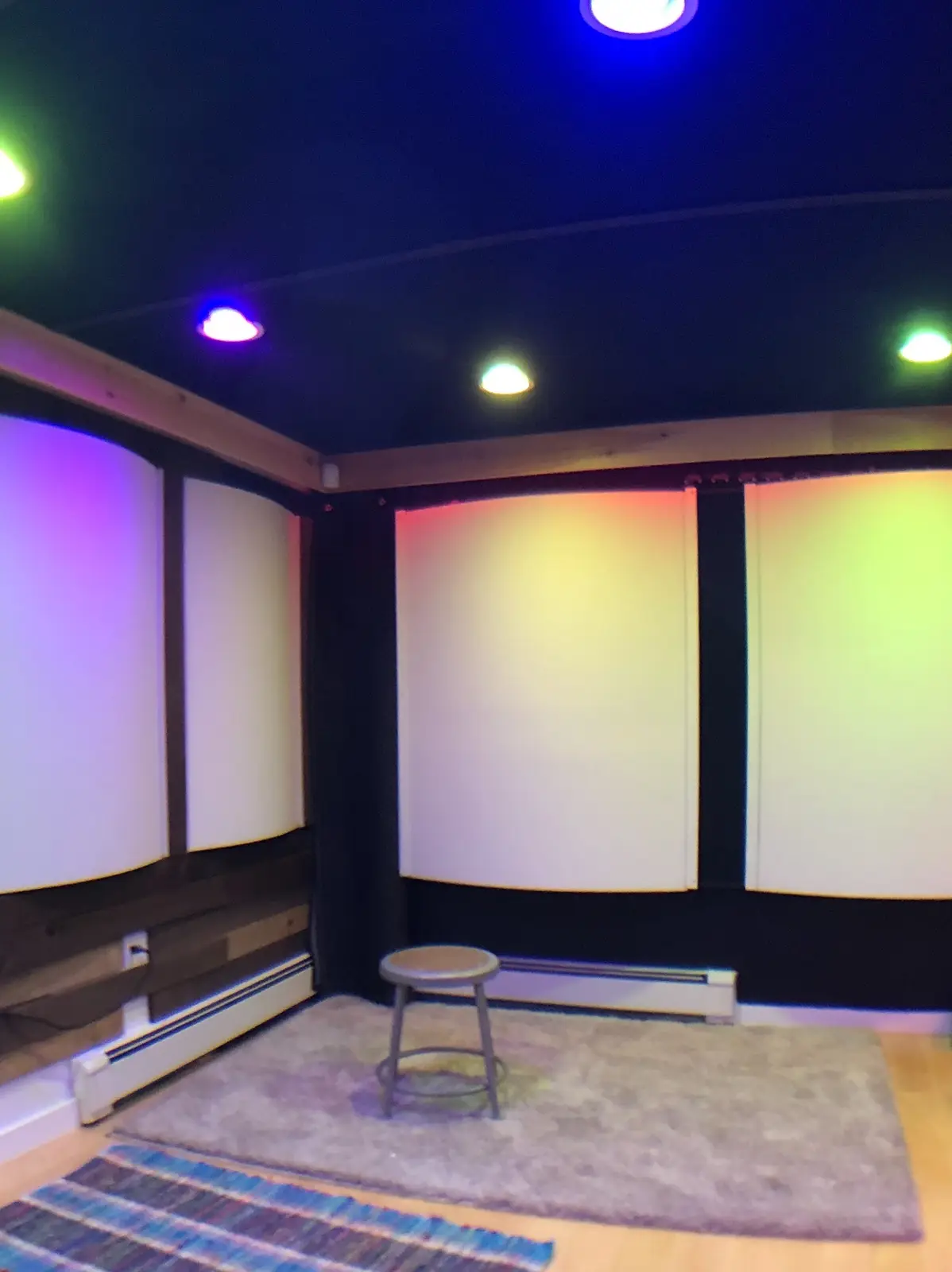 An empty home theater room with curved acoustic panels on the walls, a single stool in the center, and colorful LED lights…