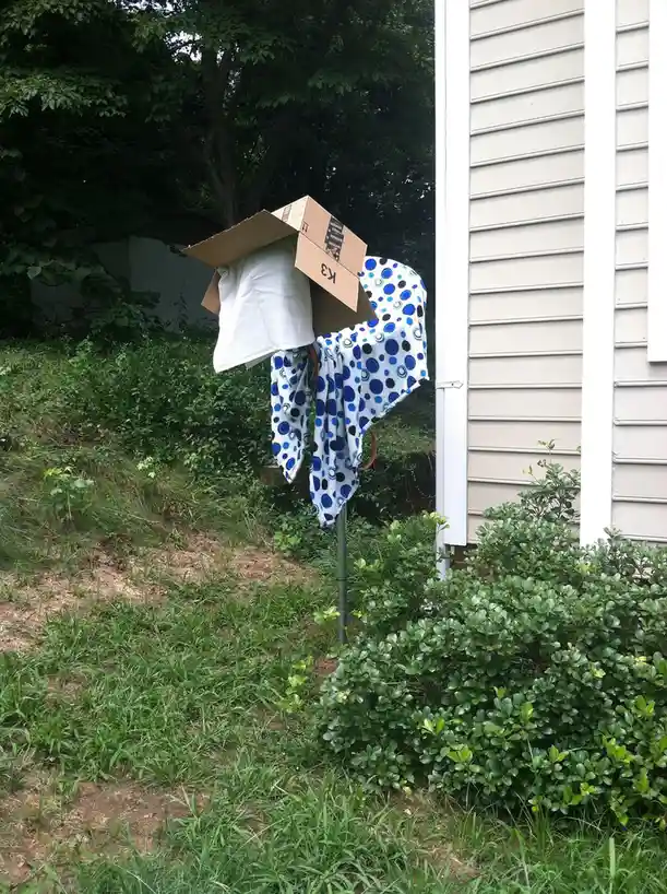 An outdoor scene shows a makeshift scarecrow or costume made from a cardboard box, a white t-shirt, and a blue-and-white…