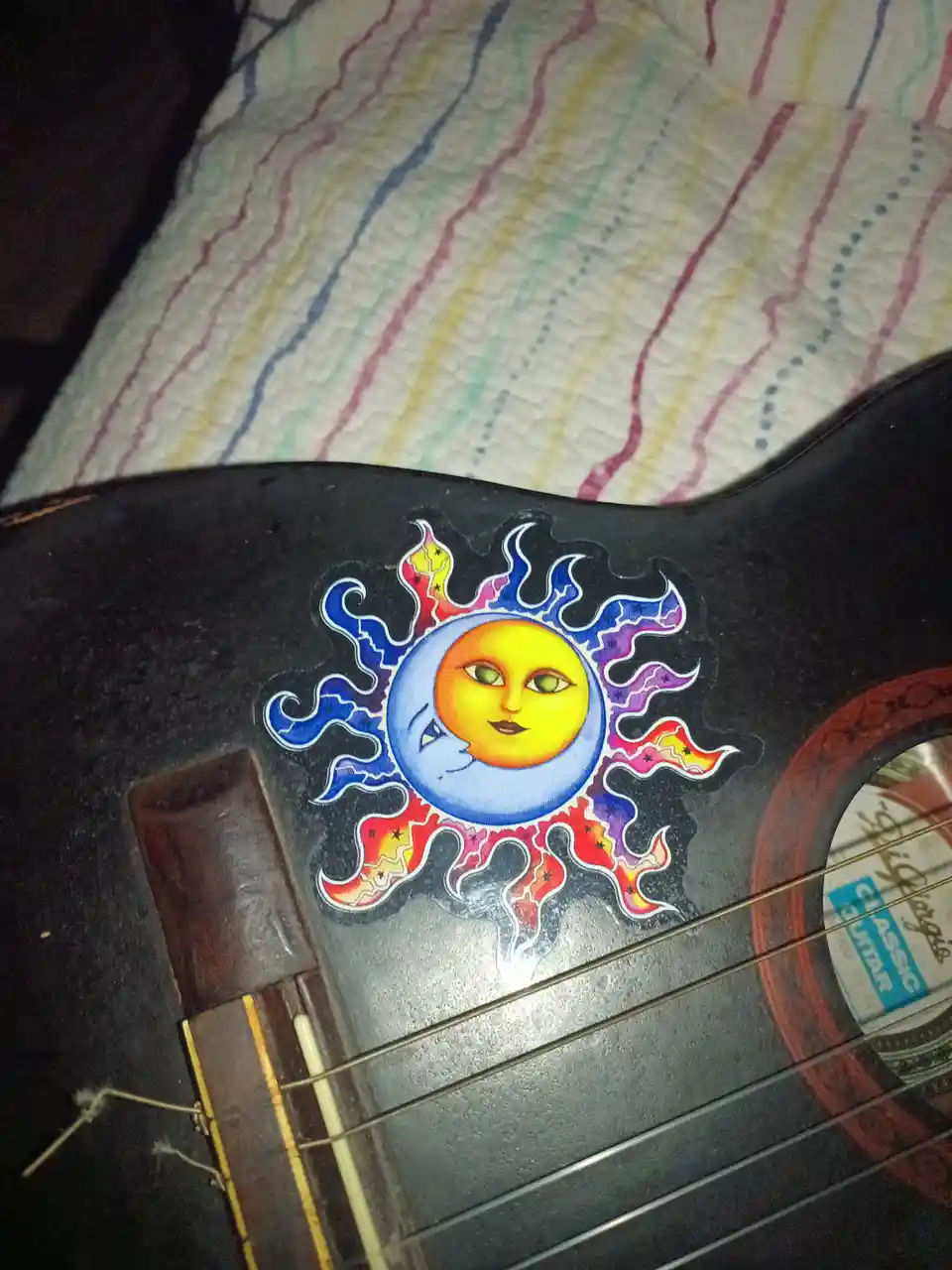 A close-up photo shows a colorful sun-and-moon sticker with a face, placed on the dark body of a classical guitar resting on…