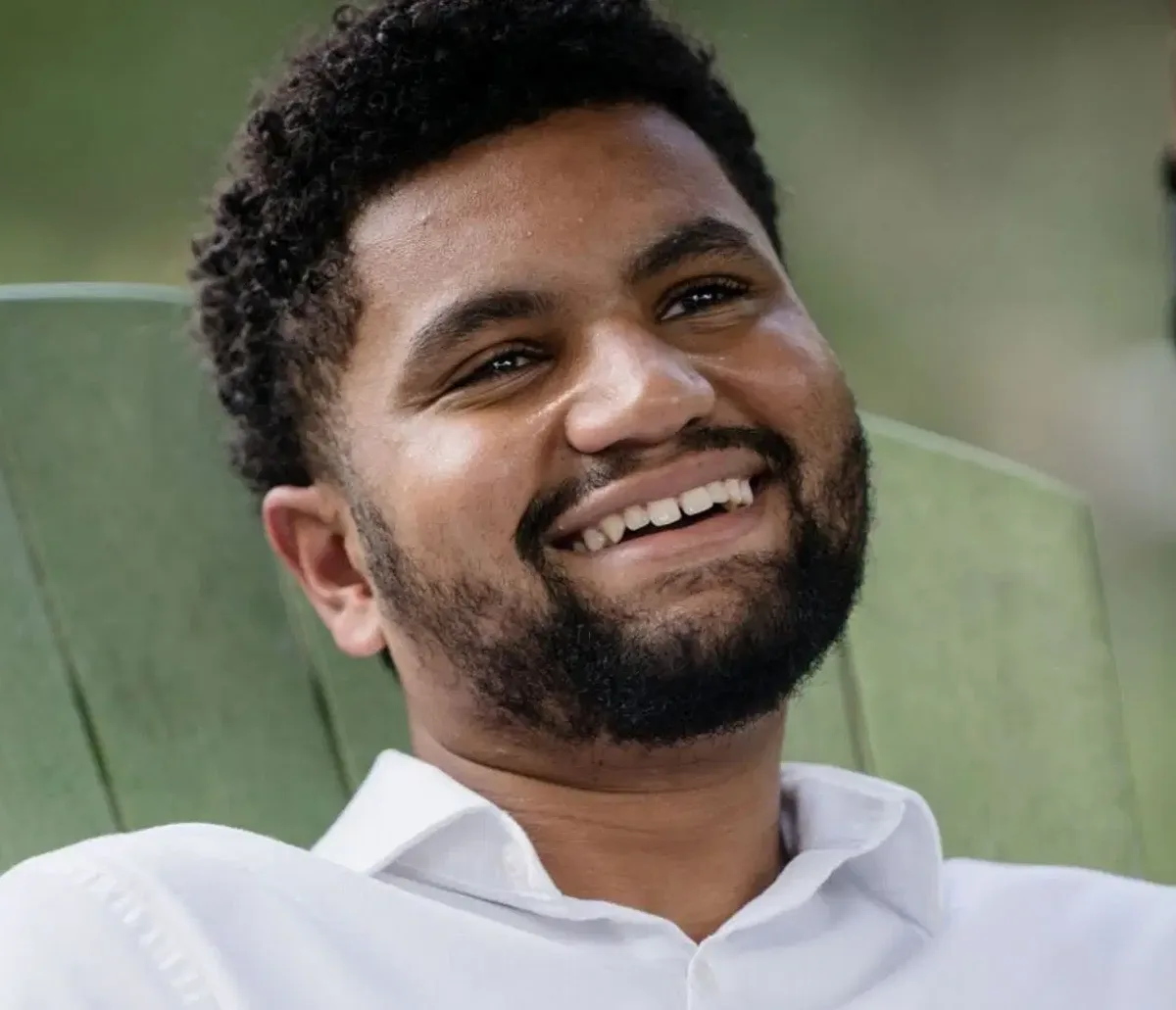 a man with curly black hair and a beard, wearing a white collared shirt, is smiling broadly while seated in a green chair…