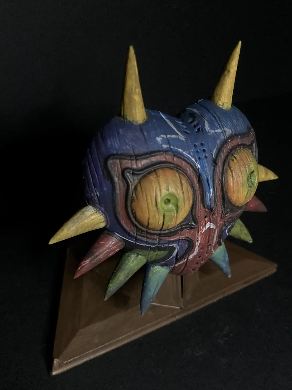 A handmade, colorful wooden mask with large, round eyes and multiple spikes in yellow, red, green, and blue, displayed on a…