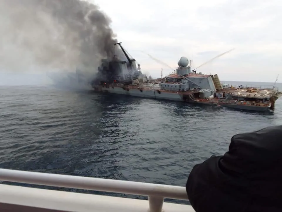 A large naval ship is on fire, emitting thick black smoke, while another vessel sprays water to extinguish the flames. In…