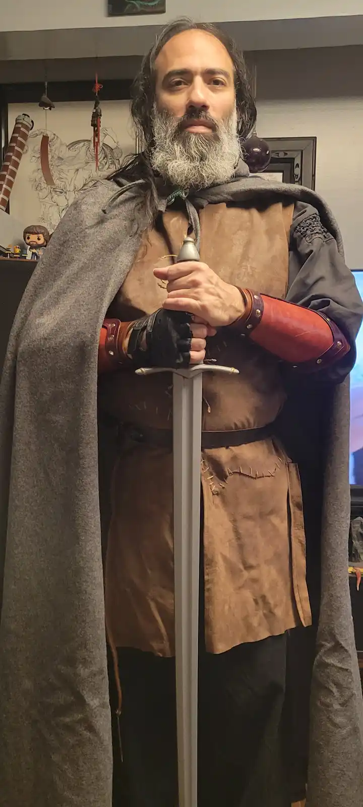 A man with a long grey beard and dark hair stands indoors wearing a grey cloak, brown leather tunic, and red leather…