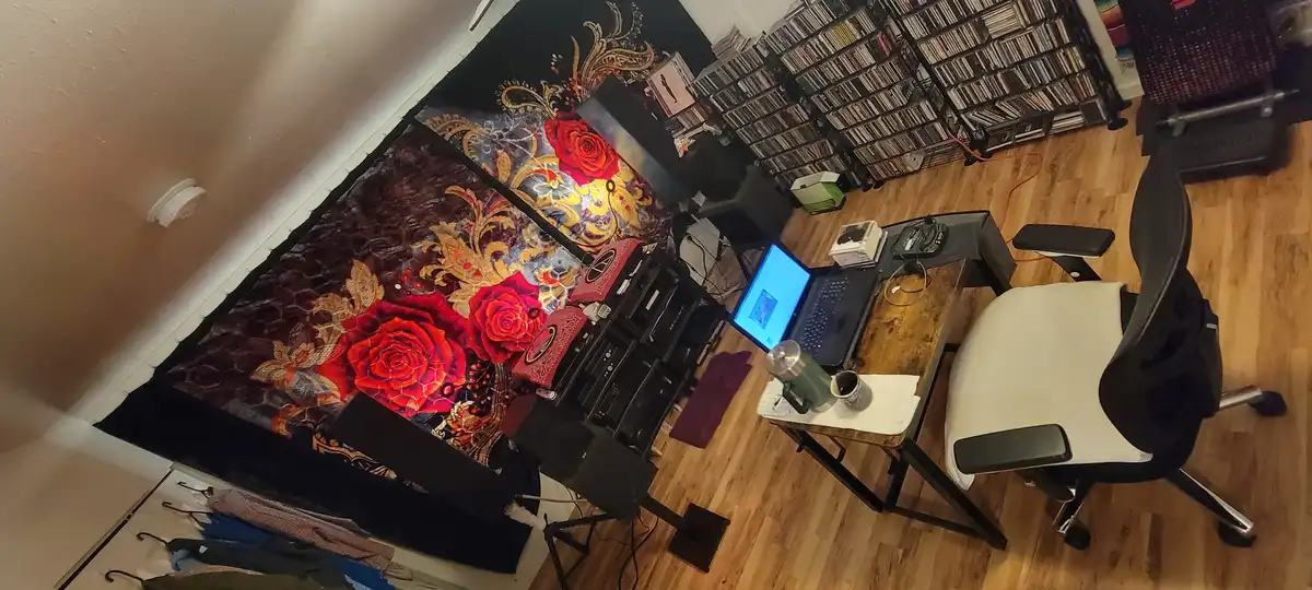 An overhead, rotated photo of a room with a desk, chair, laptop, and extensive wall-mounted racks filled with CDs or DVDs. A…
