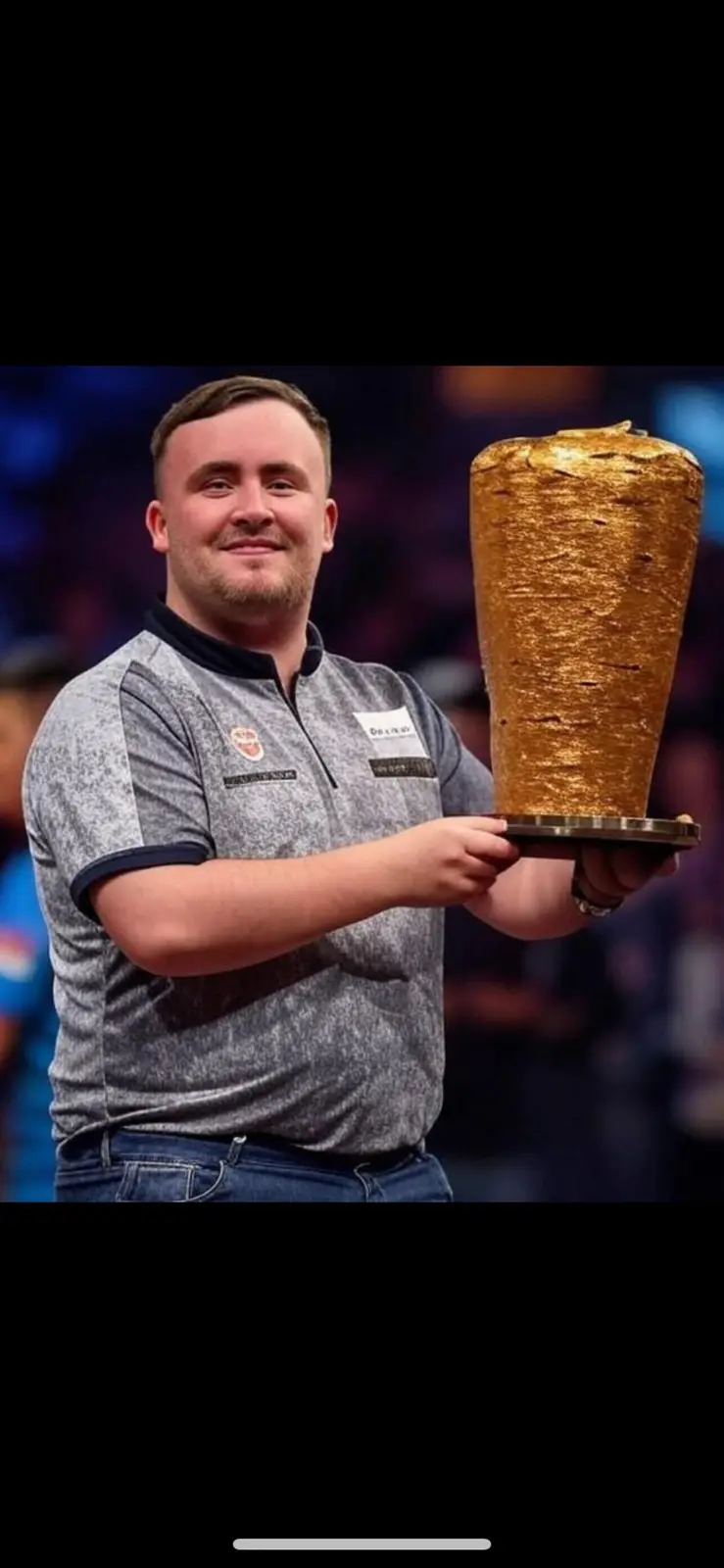 A smiling man in a grey polo shirt and jeans holds a large, golden, cone-shaped trophy that resembles a shawarma. The trophy…