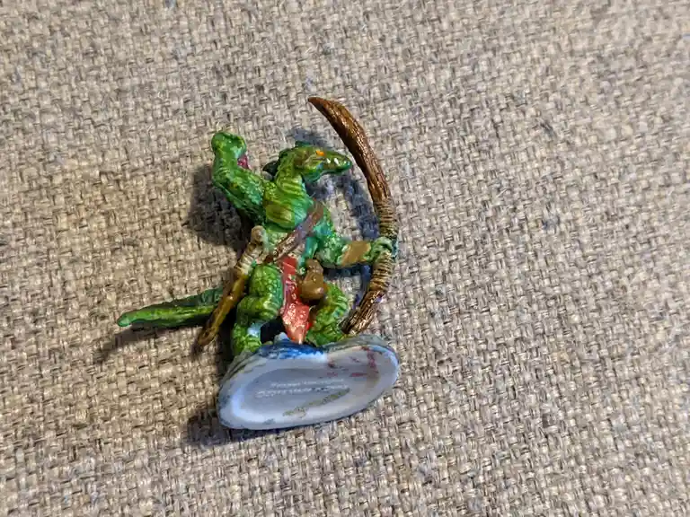 A small green plastic dragon figurine, painted with red accents and holding a curved bow, is shown from behind on a beige…