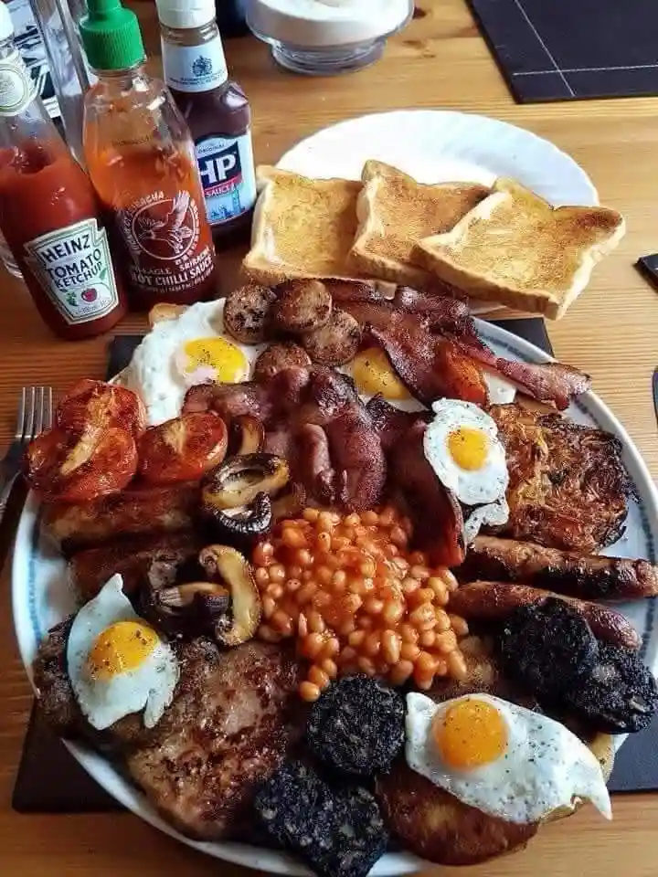 a large, overflowing plate of a traditional full english breakfast featuring fried eggs, bacon, sausages, baked beans, black…