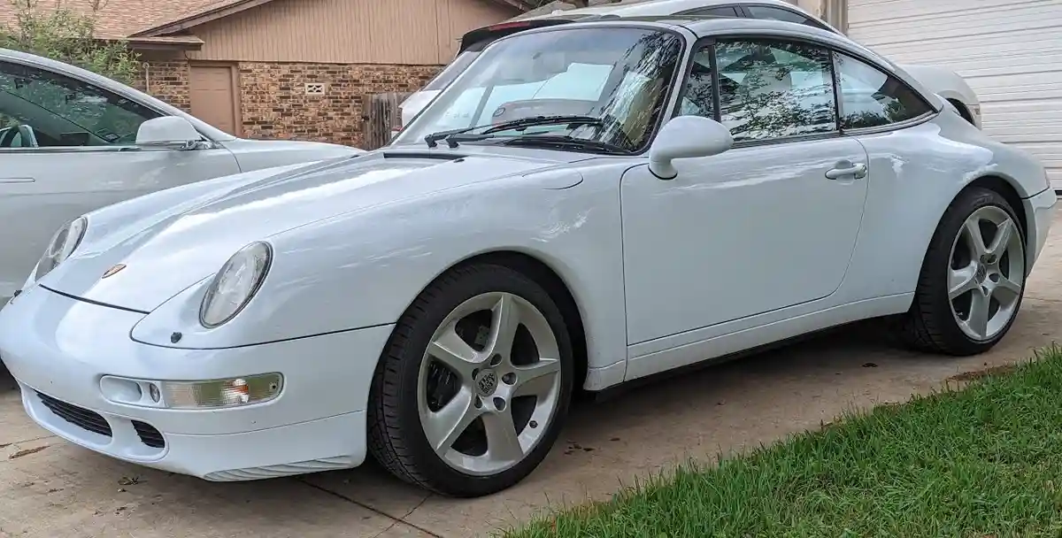 a white porsche 911 is parked on a concrete driveway next to a grassy area, with a house and another car visible in the…