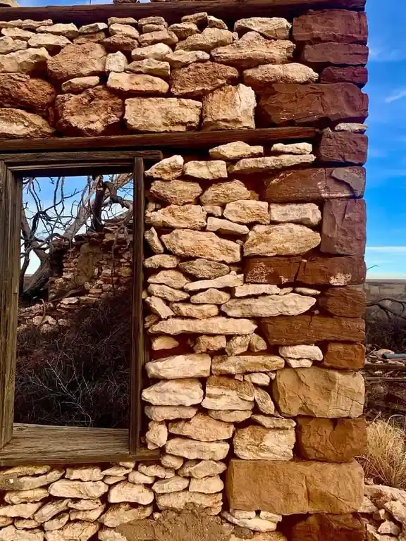 A close-up photo of a weathered stone wall with a wooden-framed window, showing the interior ruins and bare branches through…
