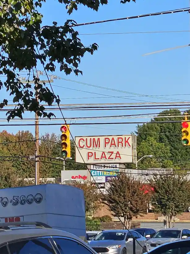 A daytime photo of a commercial plaza sign that reads 'CUM PARK PLAZA' in red letters, with traffic lights and power lines…