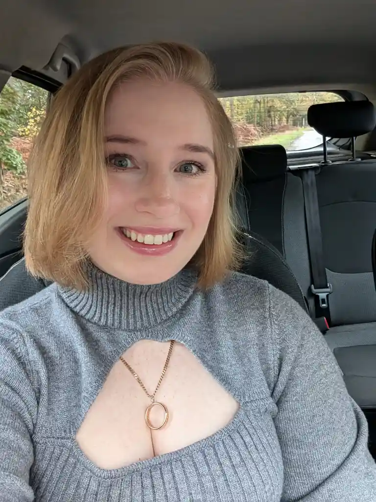 A woman with blonde hair and a grey turtleneck sweater with a heart-shaped cutout in the chest area is smiling at the camera…