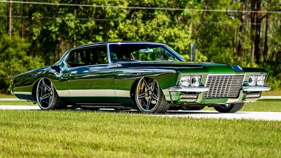 A glossy green classic American muscle car, likely a Buick Riviera, is parked on a paved surface next to grass with trees in…