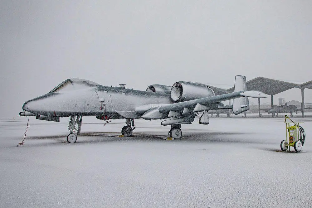 A military jet fighter, likely an A-10 Thunderbolt II, is completely covered in snow on an airfield during a heavy…