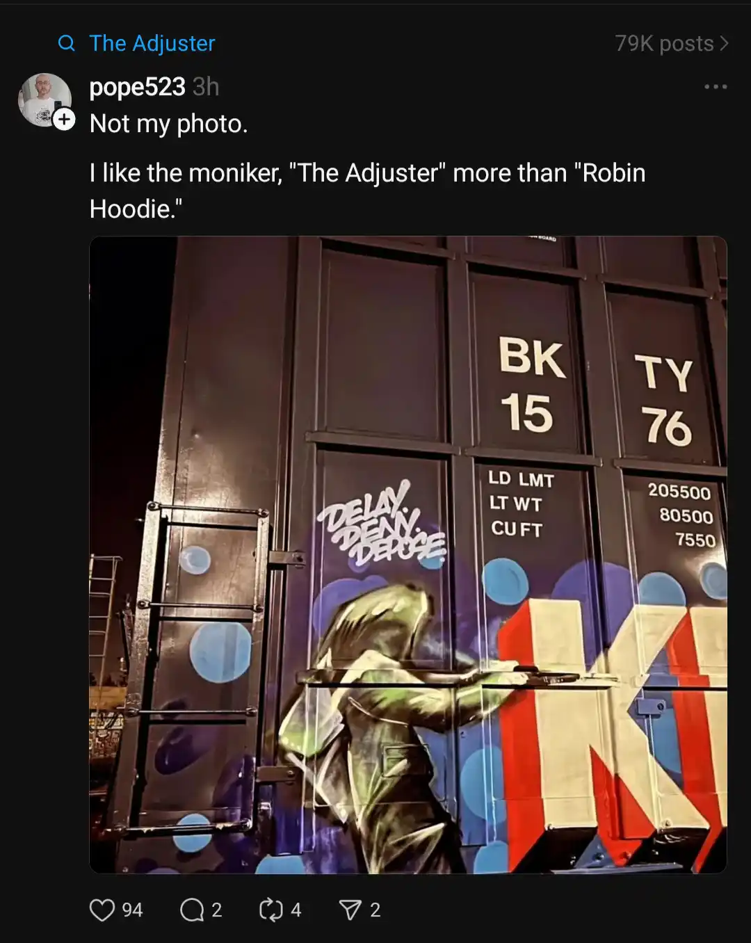 A nighttime photo of a large black shipping container with graffiti art depicting a hooded figure spray-painting a large 'K'…