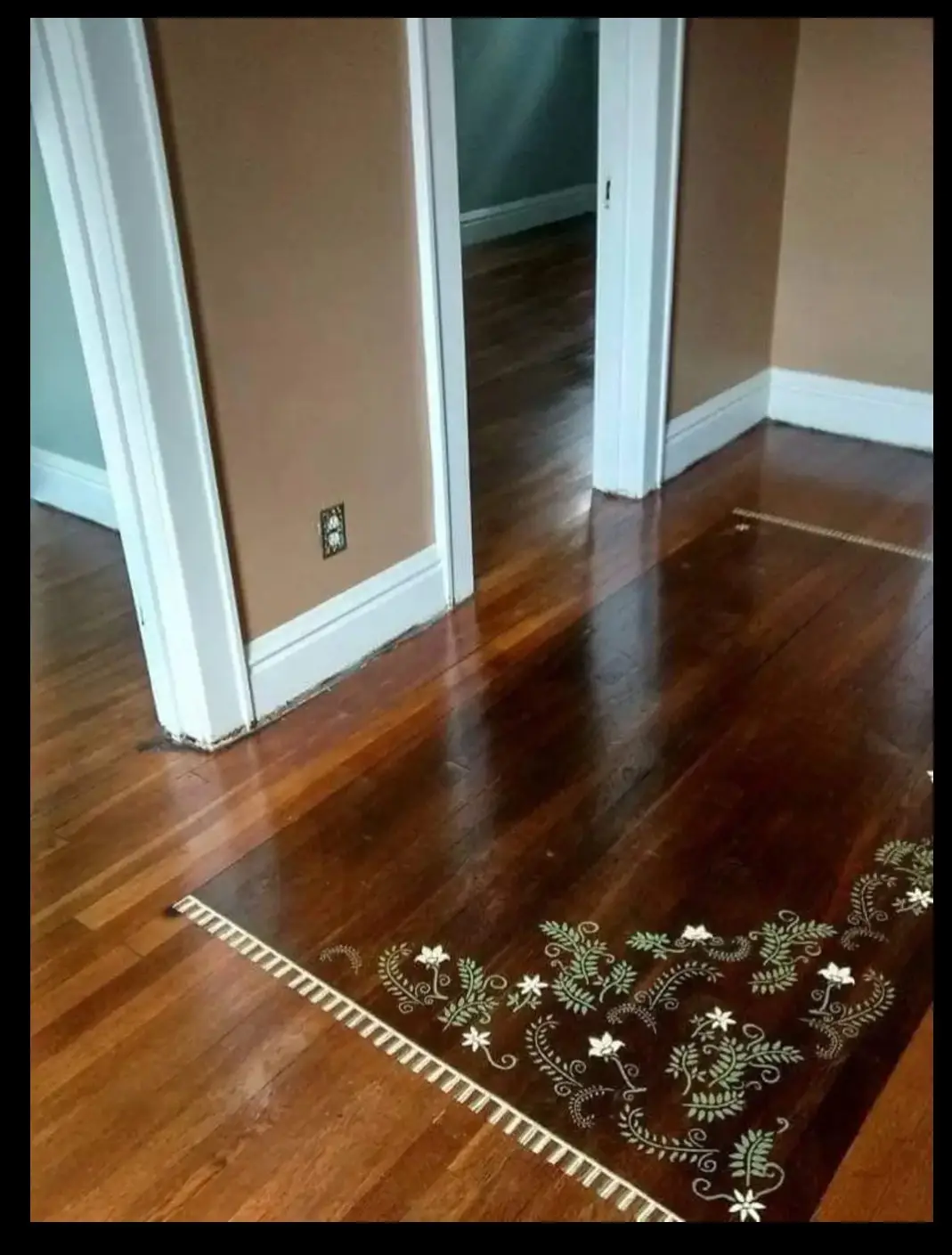 A photo shows a polished hardwood floor with a decorative inlaid border featuring floral and vine patterns in white and…