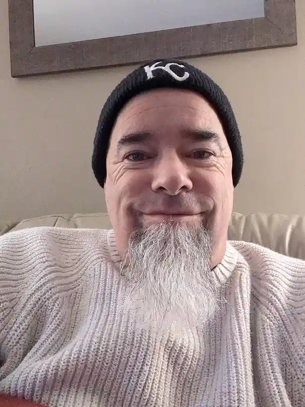 A man with a long white beard and a black beanie with 'KC' embroidered on it smiles at the camera while sitting on a beige…