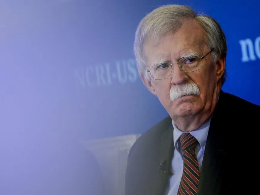 a close-up photo of an older white man with glasses and a mustache, wearing a suit and tie, looking sternly to the side…