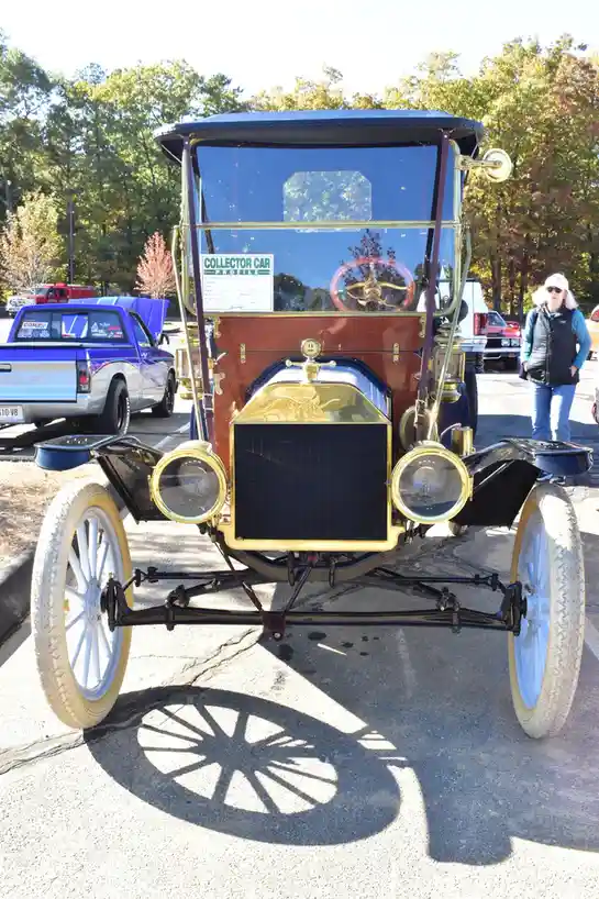 A front-facing photo of a vintage, open-top Ford Model T with a blue roof, brass accents, and white-walled tires, parked…