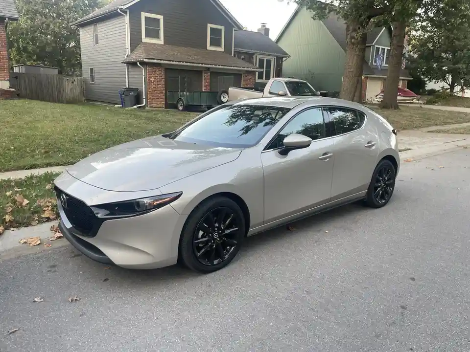 A silver Mazda 3 hatchback with black wheels is parked on a residential street in front of houses. The car is the main…