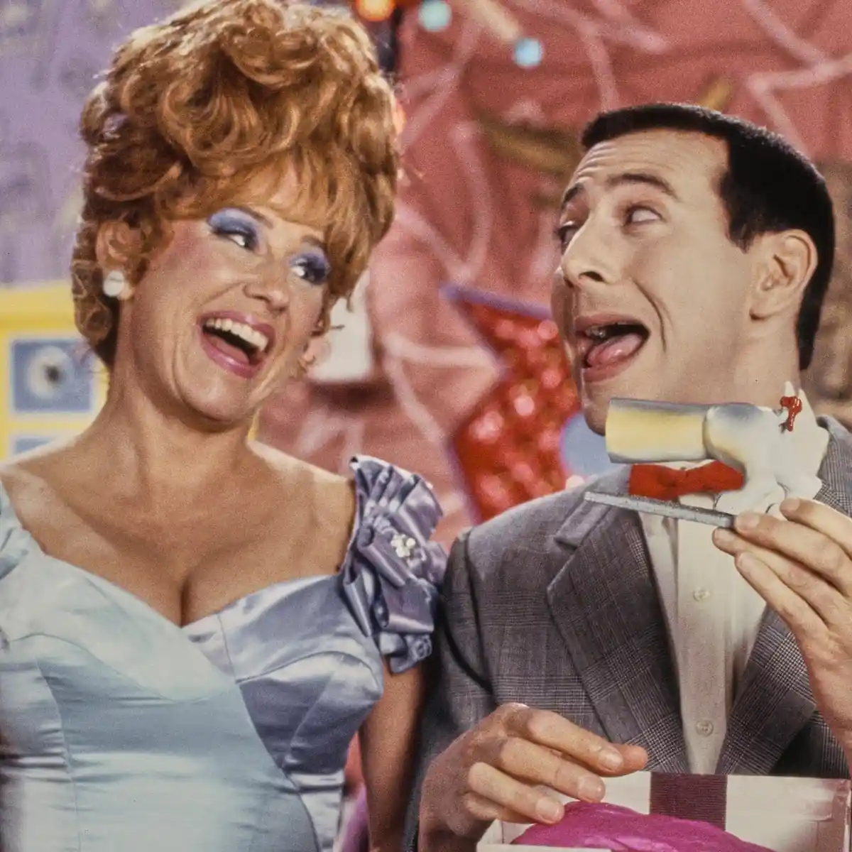 A man in a suit holds a cake with a toy gun attached to his neck, looking surprised, while a woman in a blue dress laughs…