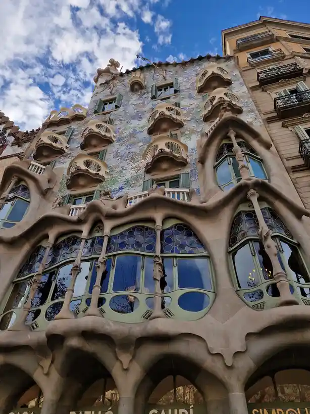 A low-angle photograph captures the ornate, organic facade of Casa Batlló in Barcelona, showcasing its bone-like balconies…