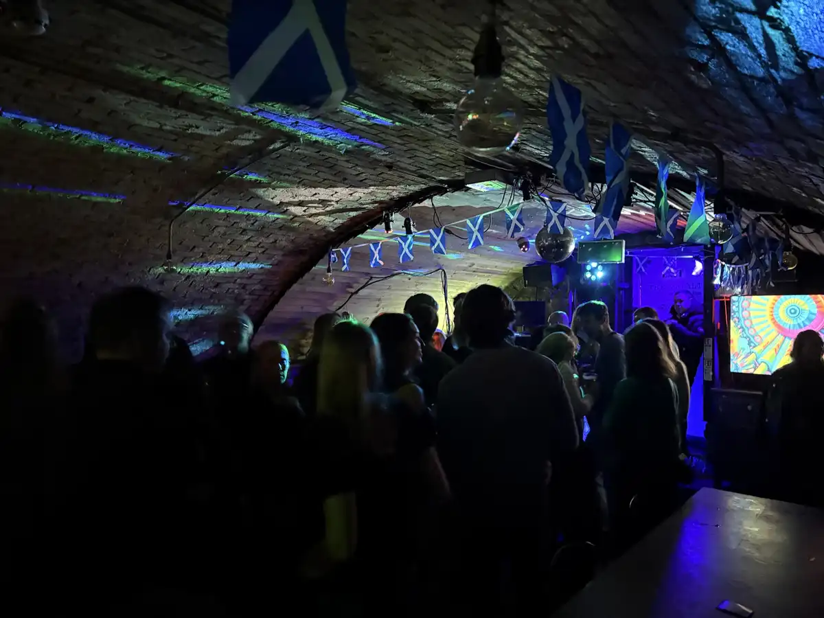 A dimly lit, crowded underground club with arched brick ceilings, decorated with numerous Scottish Saltire flags and disco…