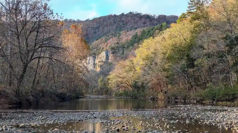 A scenic autumn river landscape with rocky shallows in the foreground, surrounded by trees with yellow, orange, and green…