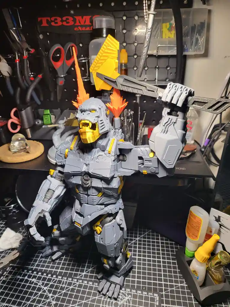 A large, grey and yellow robotic gorilla figure stands on a black grid cutting mat in a cluttered workshop, holding a large…