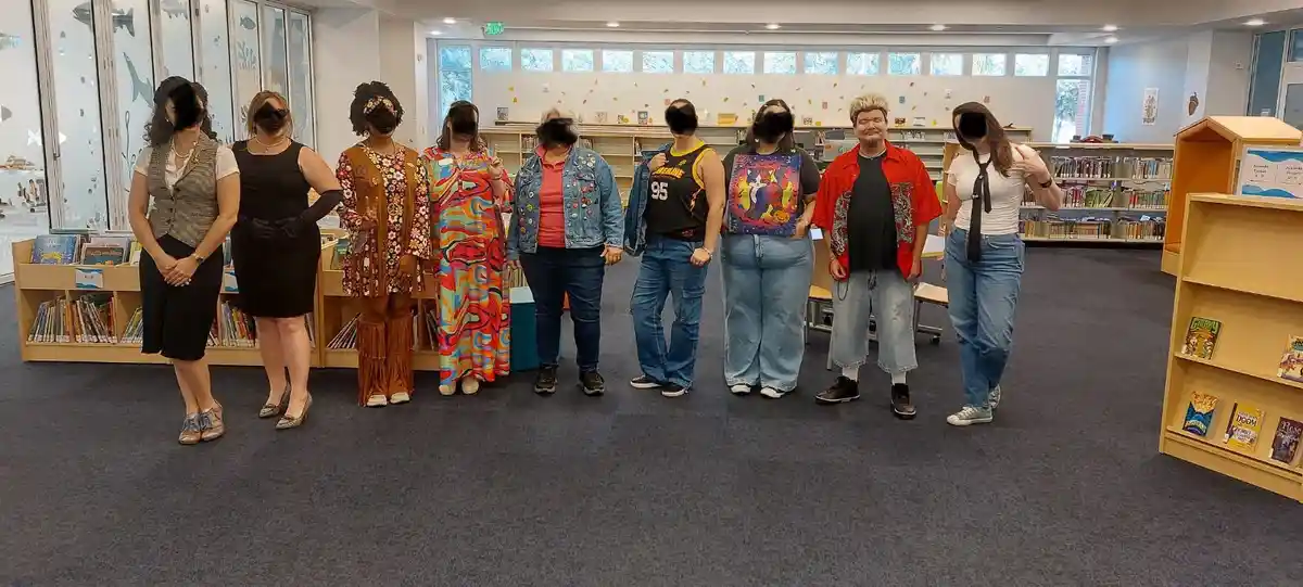 A group of eight people stand in a line inside a library, each wearing different, colorful, and eclectic outfits ranging…