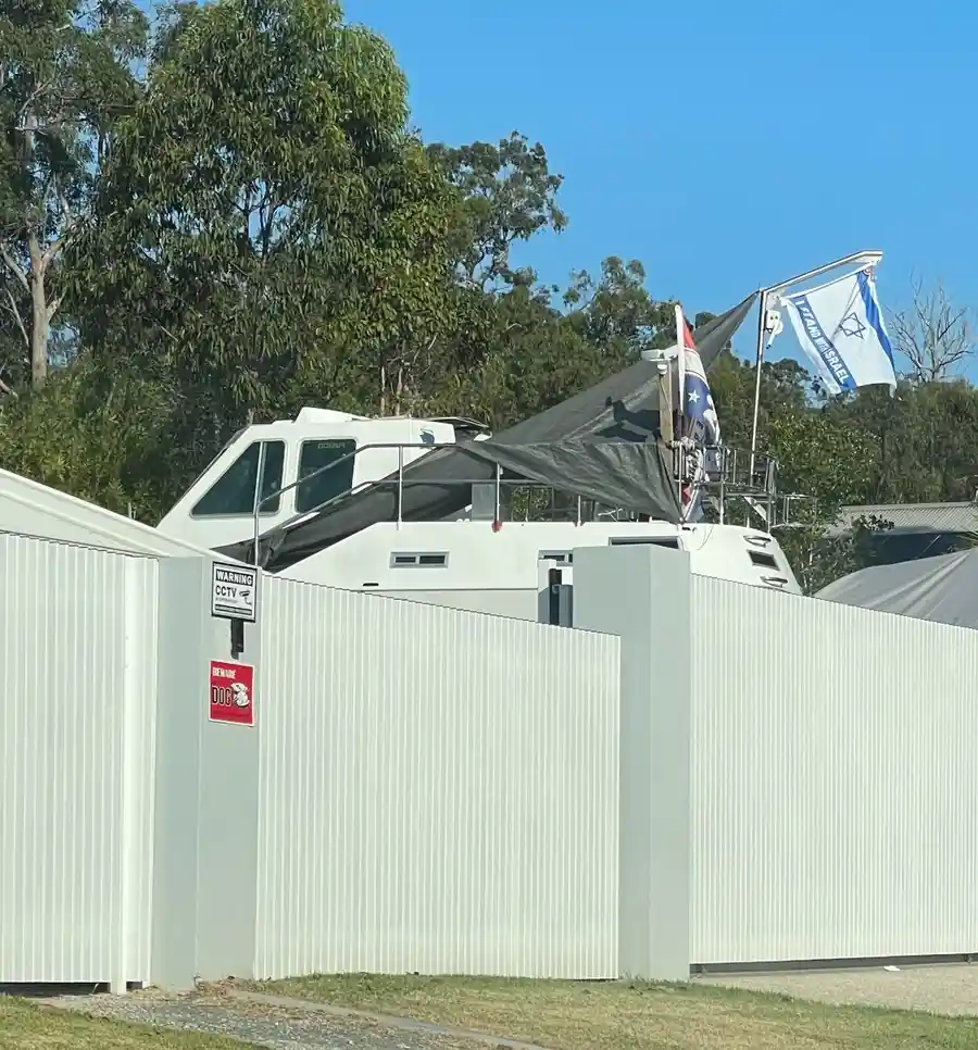 A white corrugated metal fence with a 'WARNING CCTV' sign and a 'BEWARE DOG' sticker. Behind it, a white boat-like structure…