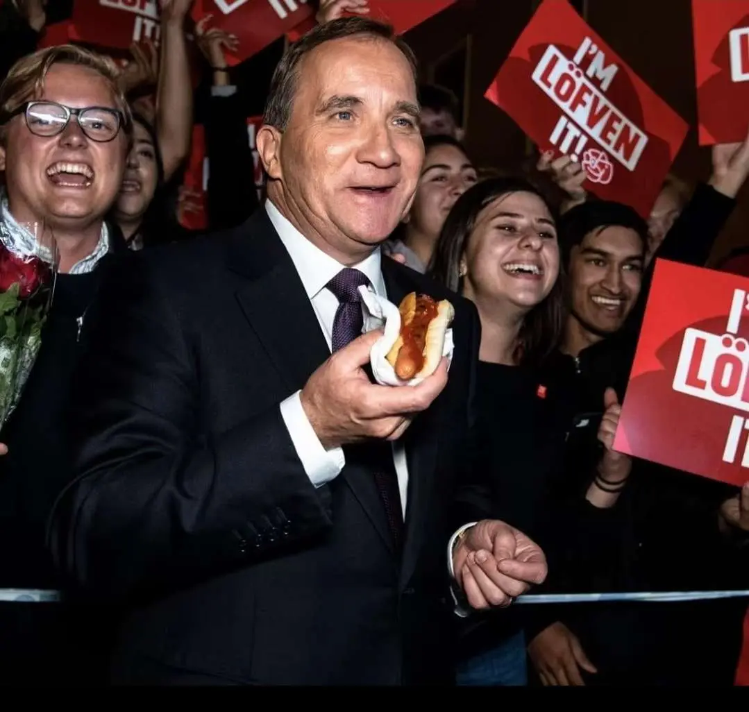 A man in a dark suit and tie holds a hot dog while smiling, surrounded by cheering supporters holding red signs that read…