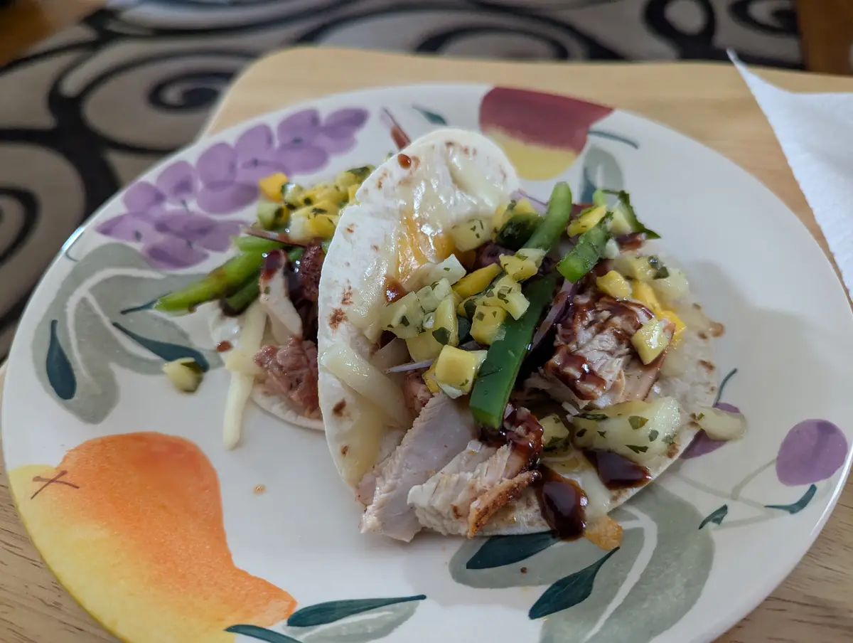 A close-up photo shows two soft tacos on a colorful floral plate, filled with shredded chicken, melted cheese, diced mango…