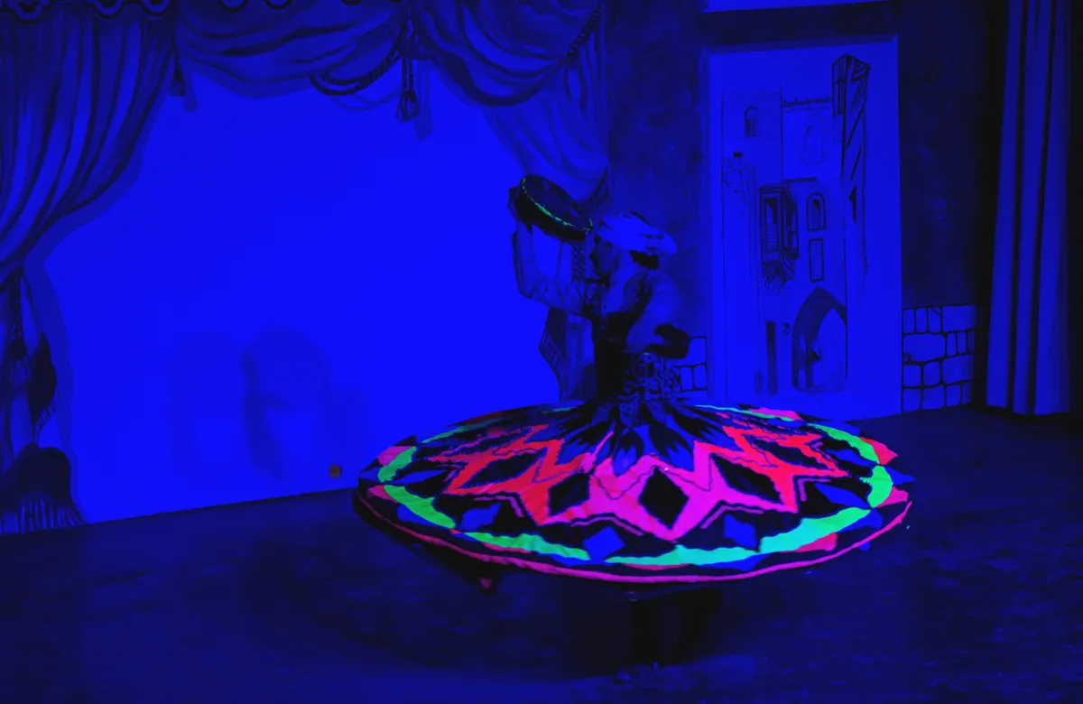A dancer in a brightly colored, glowing costume spins on a stage under blue lighting, holding a tambourine. The background…