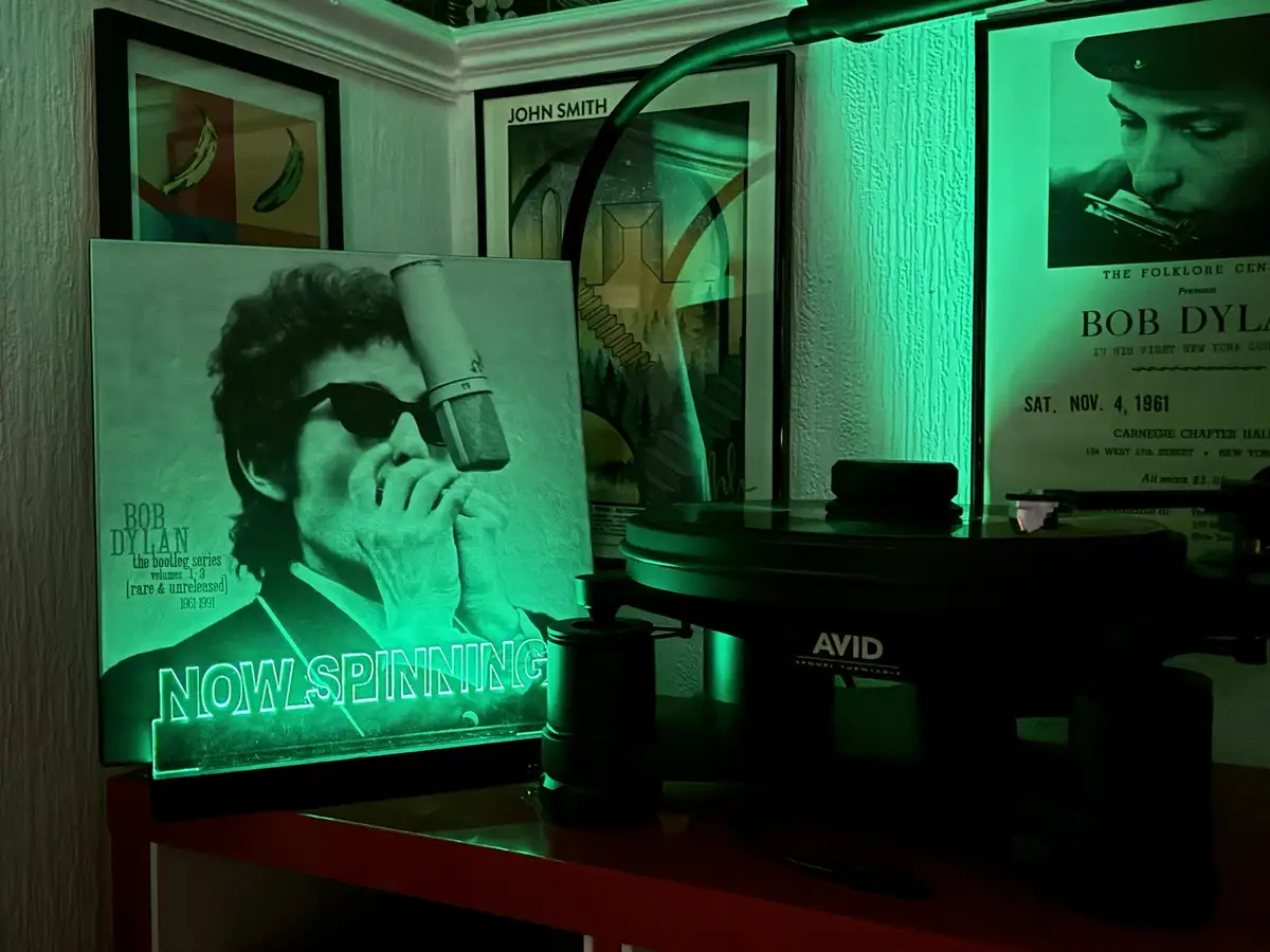 A dimly lit room bathed in green light features a record player with a Bob Dylan album cover prominently displayed. The…