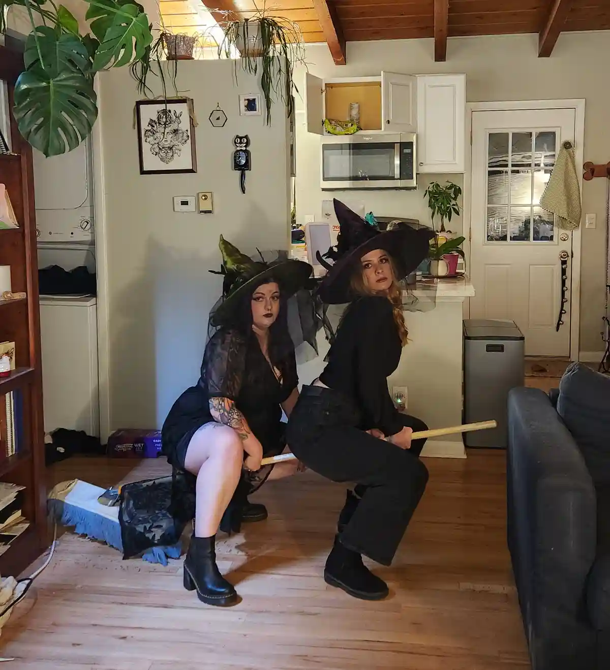 Two women dressed as witches in black costumes and pointed hats are squatting indoors on a wooden floor, each holding a…