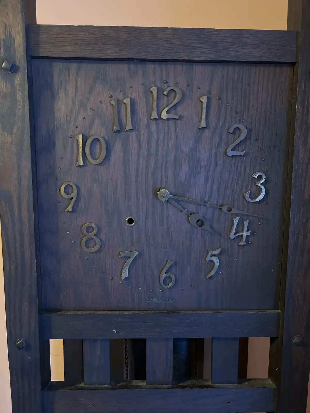 A dark wooden wall clock with metal numerals shows the time as 4:00, with the hour and minute hands both pointing to 4. The…